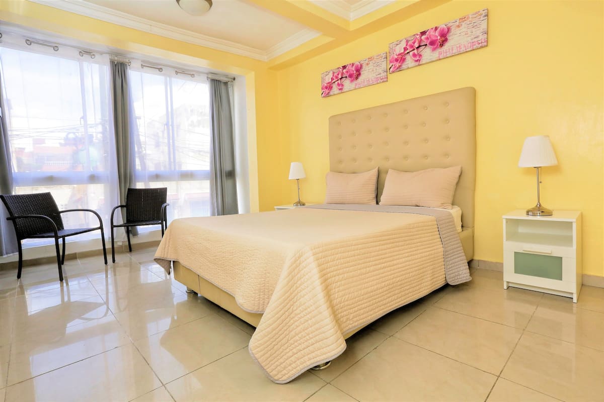 A hotel room with a large bed, two chairs, a nightstand, and a window with curtains.