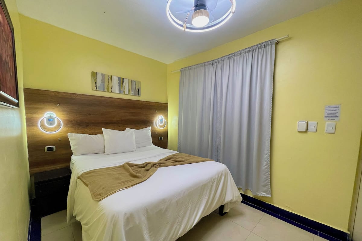 A hotel room with a bed, a wooden headboard, a ceiling fan, and a window with curtains.