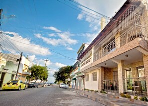A street view with a two-story building featuring a balcony with metal railings, a visible number '56', and a small outdoor seating area.