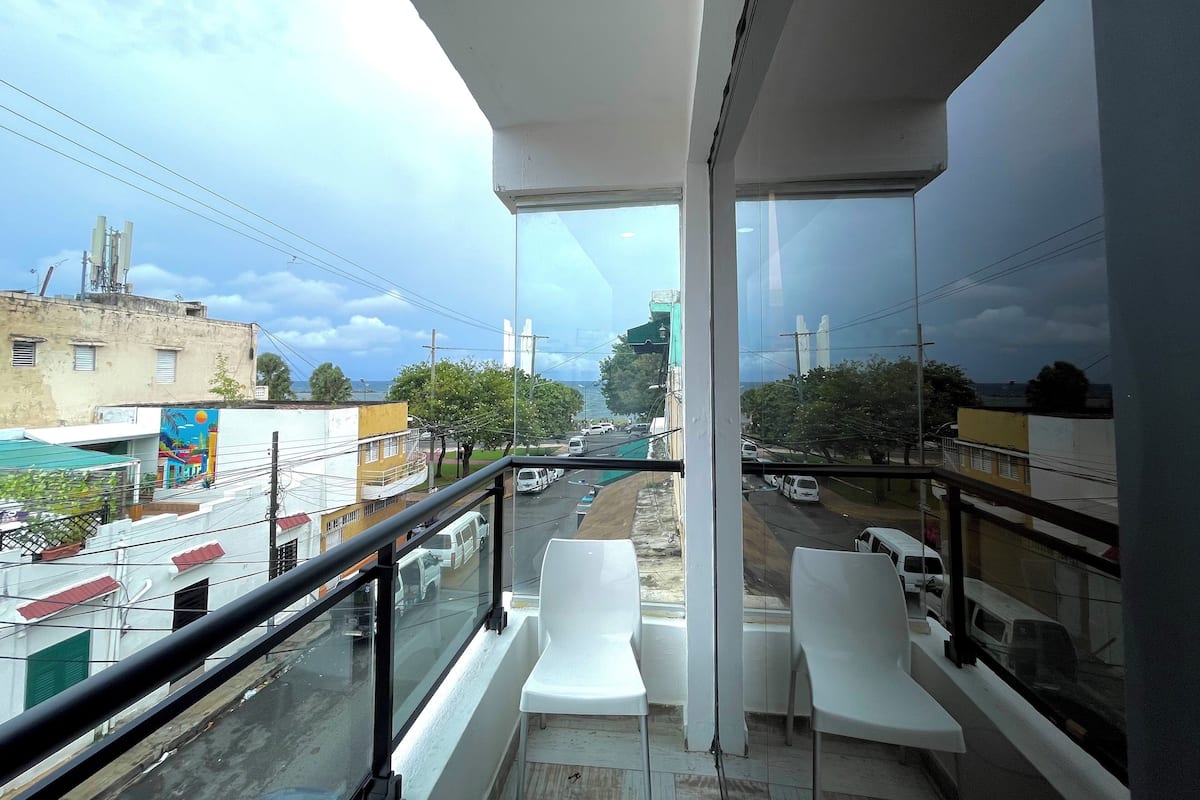 A balcony with white chairs, a glass railing, and a view of a street with buildings and cars.