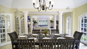A dining area with a table set for six, surrounded by wooden chairs, a chandelier, and large windows. - Green Monkey Villa (Holetown)