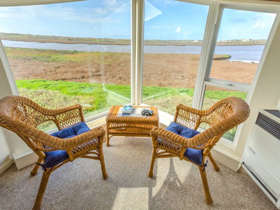 Una habitación con dos sillas de mimbre y una mesita, ventanas amplias con vista a una playa y al agua.