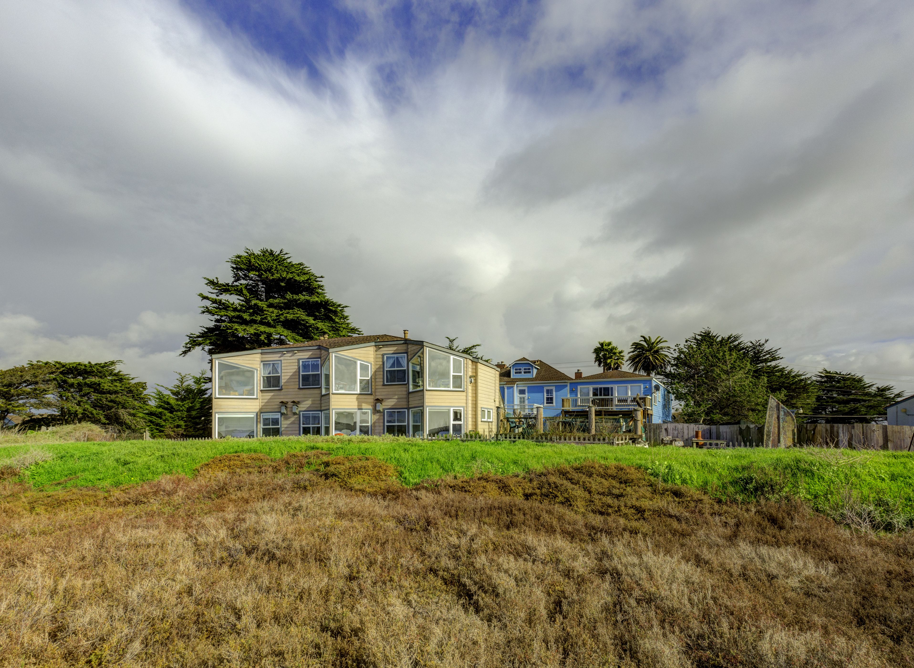 A two-story house with large windows and a sloped roof, surrounded by a grassy field and trees.