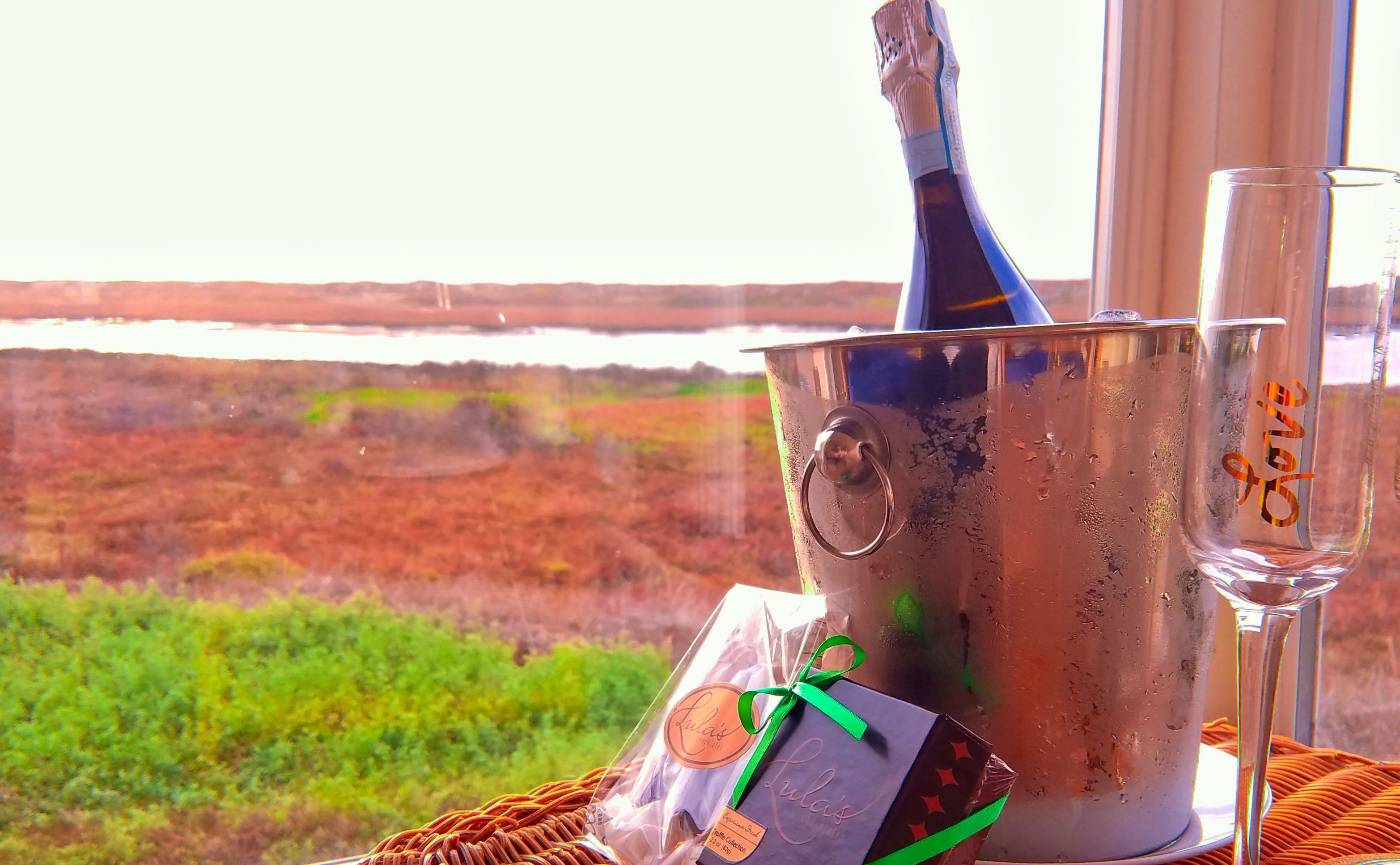 A bottle of champagne, a glass, and a gift bag with a ribbon, set on a table with a view of a landscape through a window.