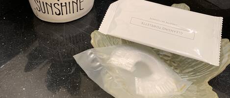 Ruukussa oleva mehikasvi, jonka kyljessä lukee teksti ”YOUAREMY SUNSHINE”, sen vieressä paketti ”CLEANSING TOWEL” -puhdistuspyyhkeitä ja heijastavalla pinnalla muovipussi.