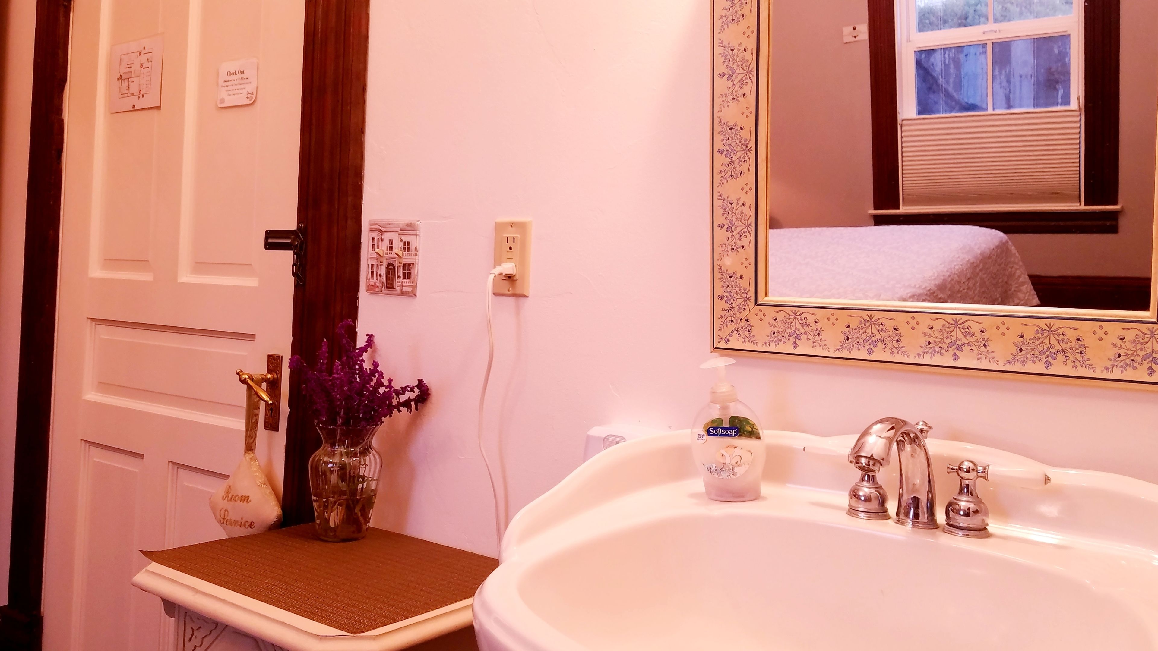 Une salle de bain avec un lavabo blanc, un miroir orné d’un cadre floral, un vase contenant des fleurs violettes et un porte-serviettes.