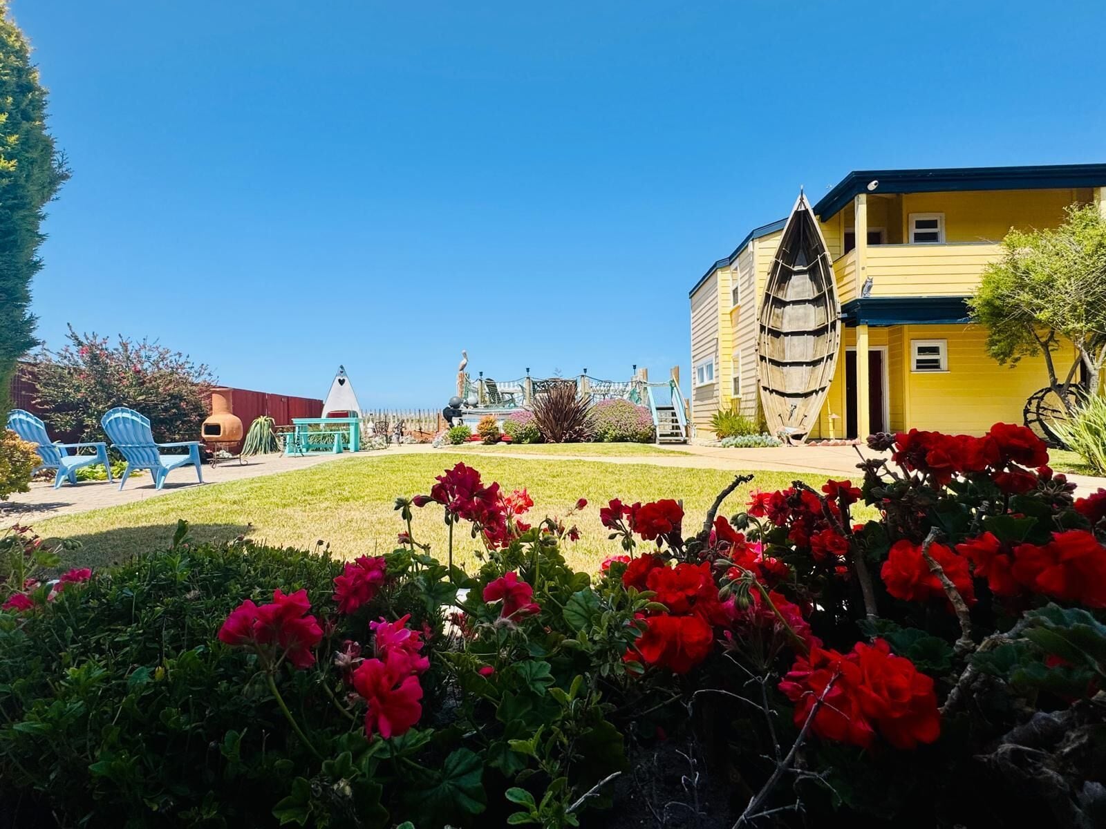 Un edificio amarillo con una gran estructura de rueda de madera en el lateral, un parque infantil con sillas azules y flores rojas en primer plano.