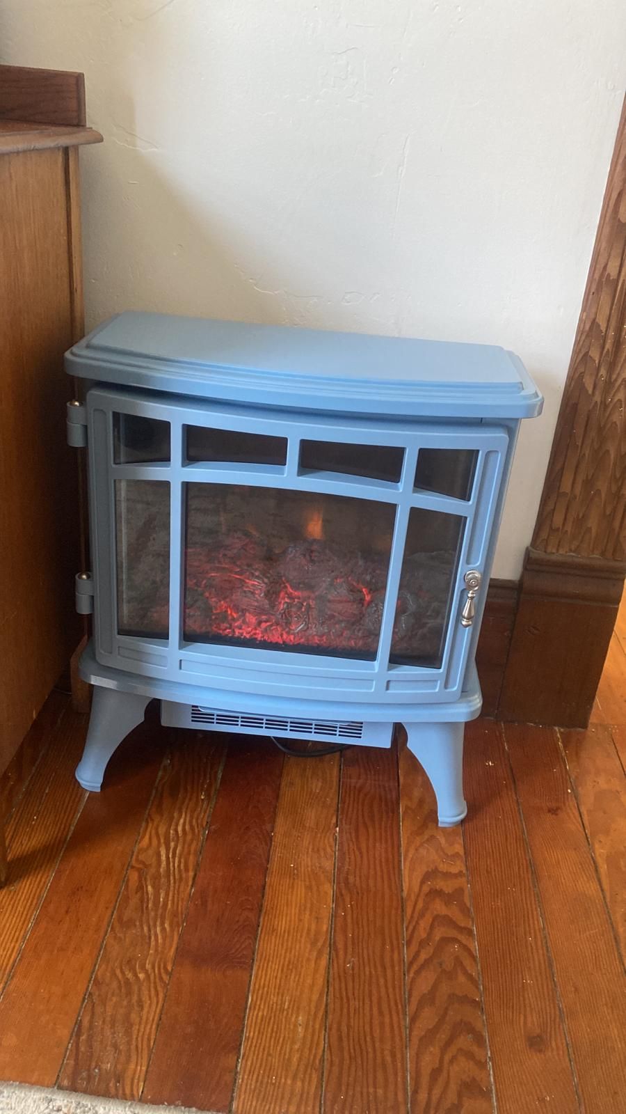 Een blauwe houtkachel met glazen deur, geplaatst op een houten vloer tegen een witte muur.