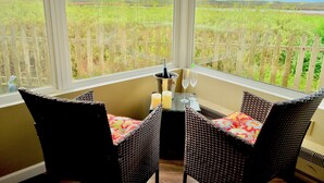A corner room with wicker chairs, a small table with candles and glasses, and a view of a fenced field through large windows. - Captain's Inn at Moss Landing (Moss Landing)
