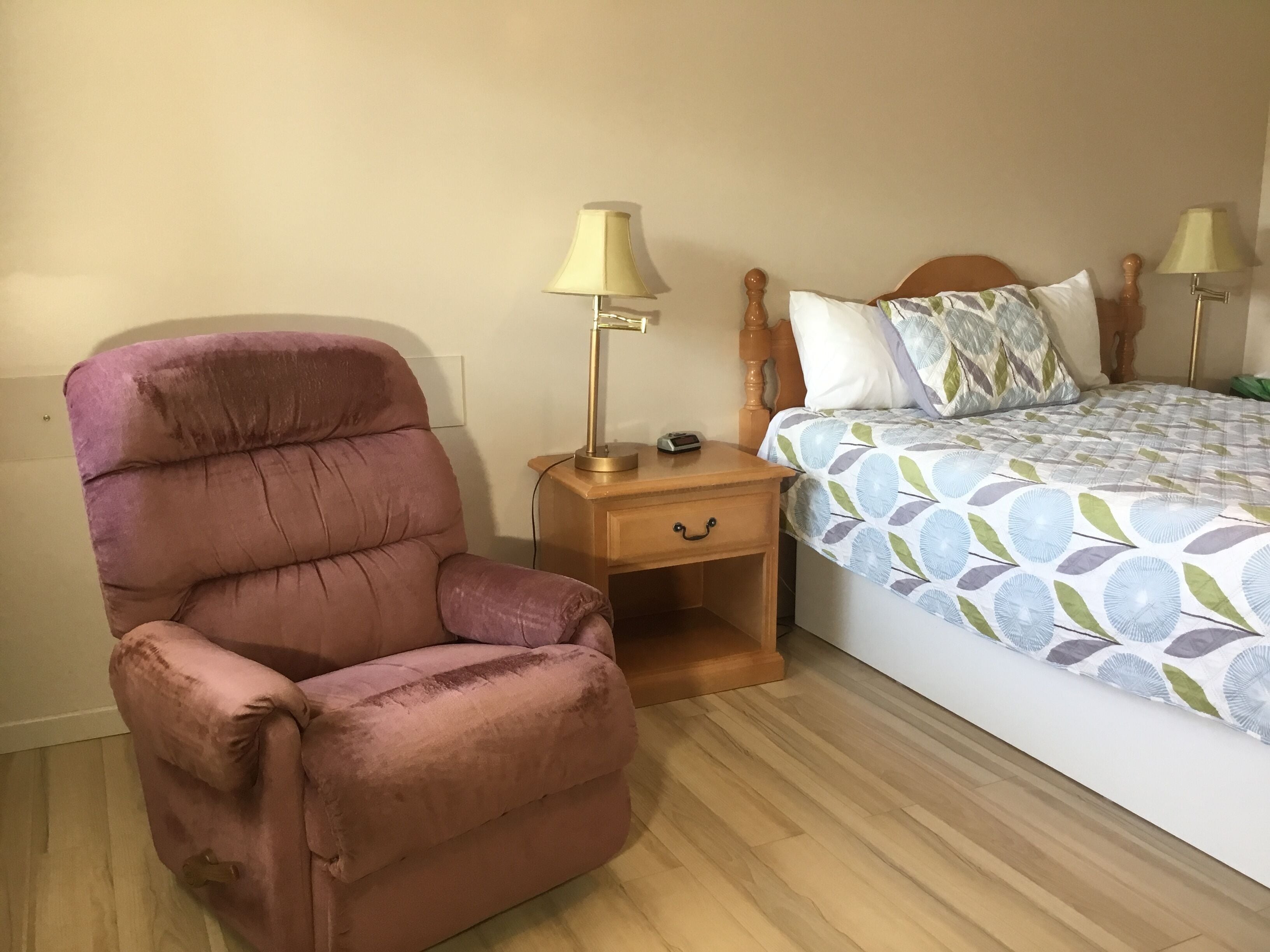 A room with a wooden bed, a wooden nightstand, a cushioned chair, and two lamps.