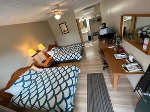 Habitación de hotel con dos camas, un escritorio, una silla, un espejo, un ventilador de techo y un baño visible a través de una puerta abierta.