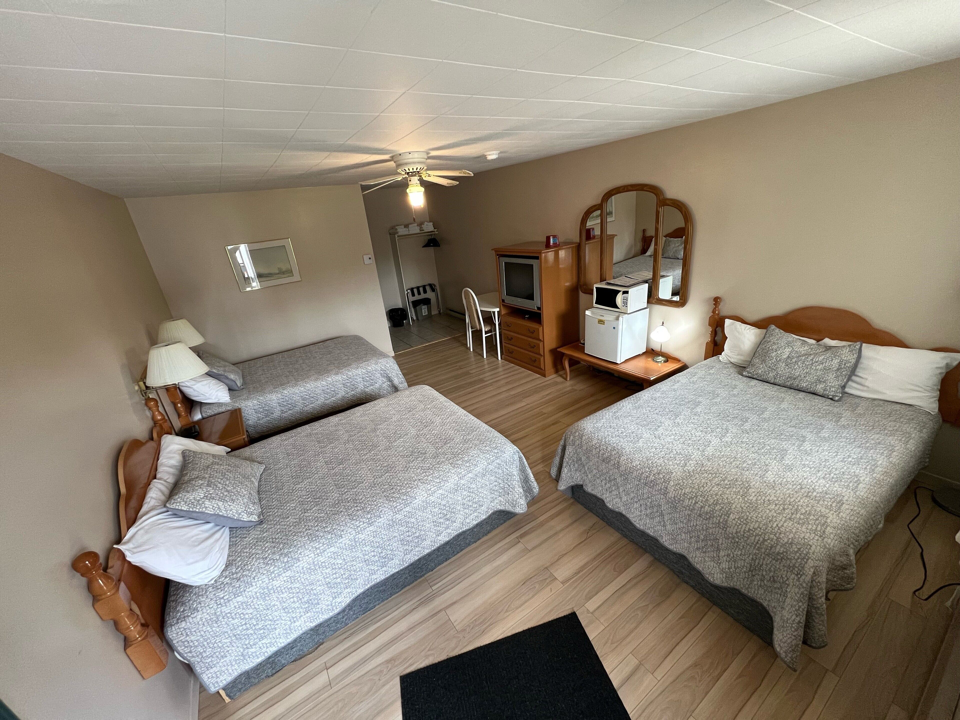 Une chambre d’hôtel avec deux lits, un bureau, une chaise, un miroir, un ventilateur de plafond et un téléviseur.