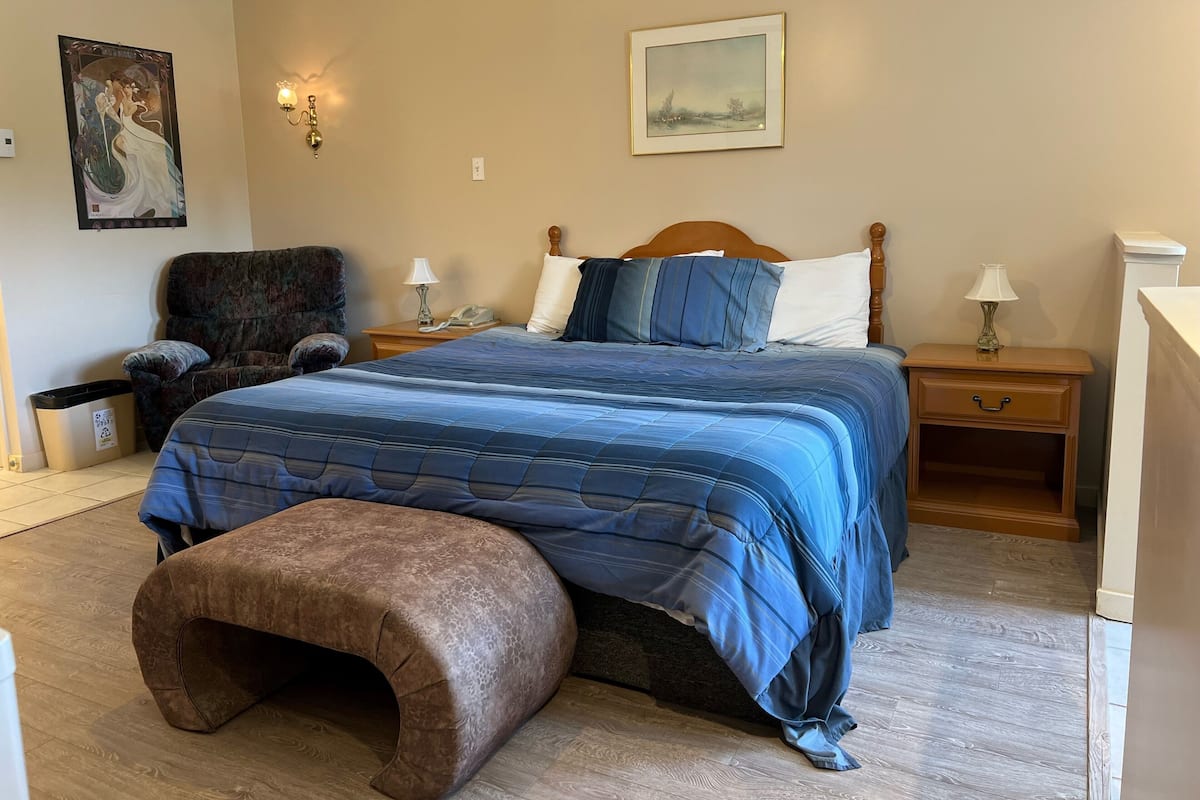 A bedroom with a large bed, a nightstand with a lamp, a chair, and a painting on the wall.