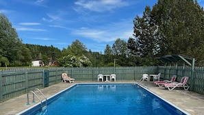Piscina rectangular con muelle de madera, rodeada por una cerca de madera, y equipada con sillas de descanso y una zona de estar cubierta.