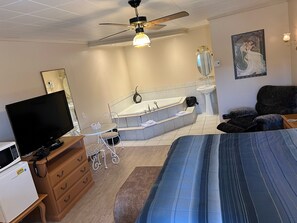 Un dormitorio con cama, televisor, escritorio, cómoda, silla, ventilador de techo, bañera y un cuadro en la pared.