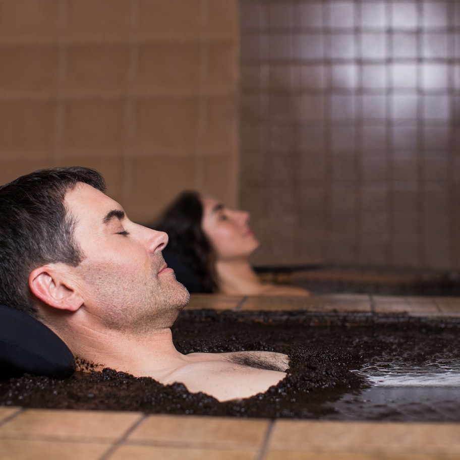 A person lying in a hot tub with a black pillow, surrounded by tiled walls.