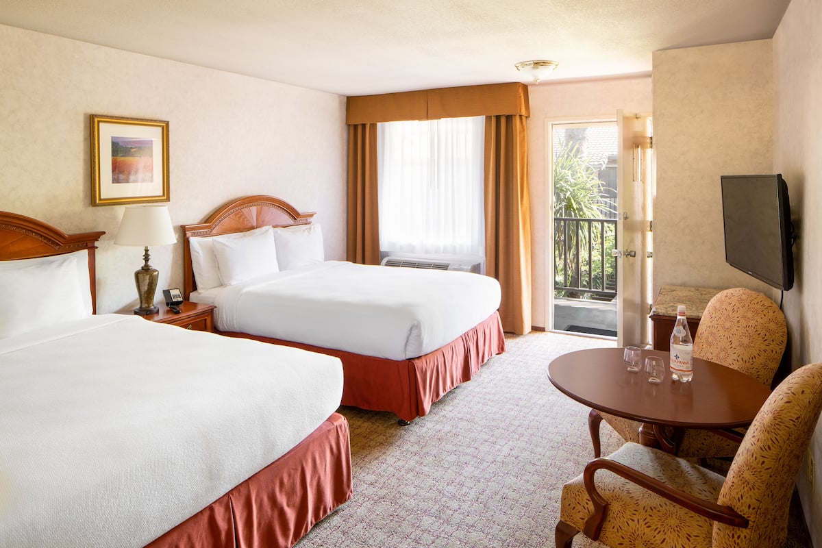 A hotel room with two beds, a small table with glasses and a bottle, a chair, a TV, and a balcony with curtains.