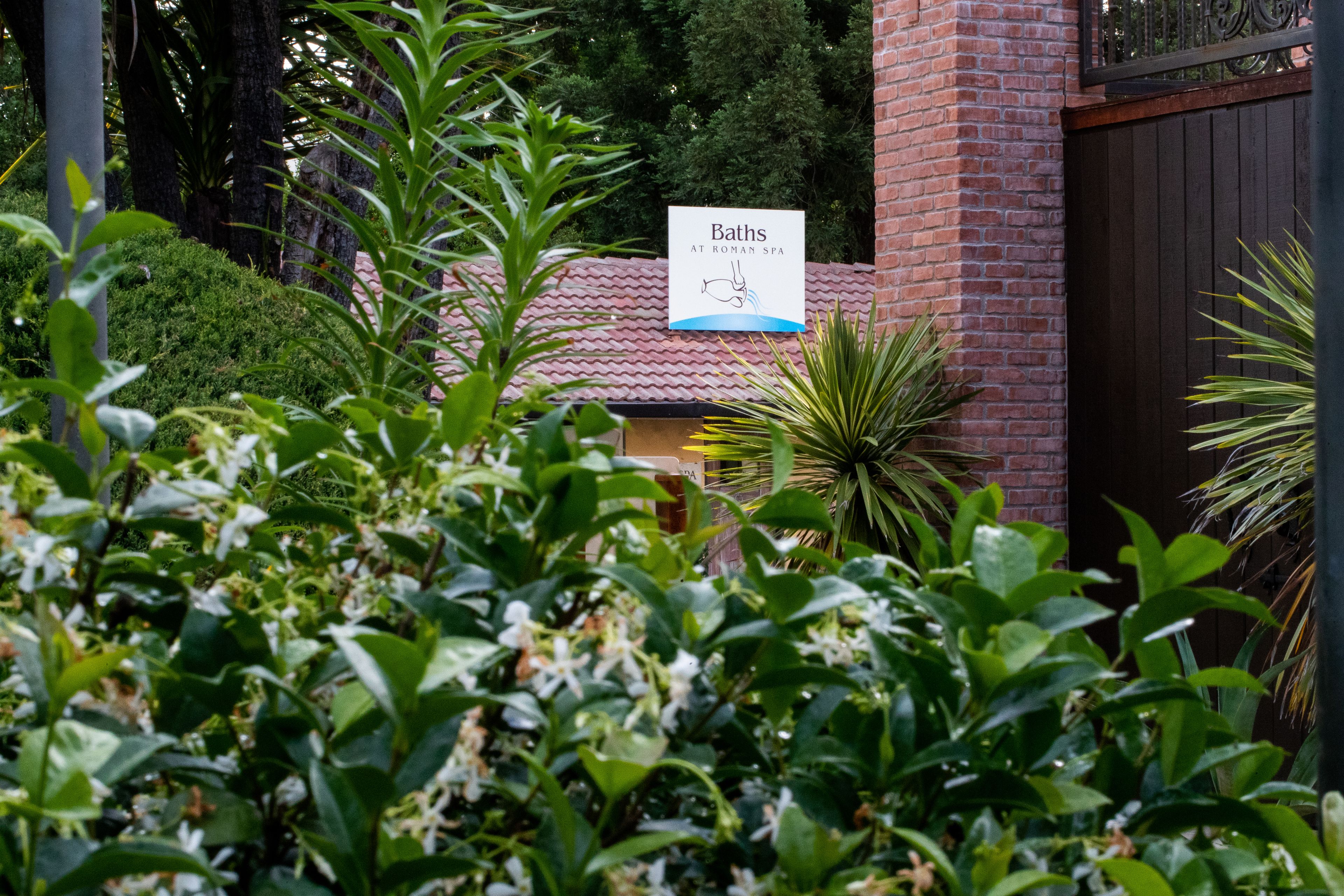 A sign for Baths at Roman Spa, surrounded by lush greenery and a brick building.