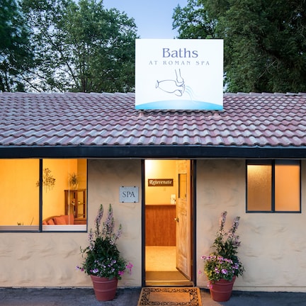 A building with a tiled roof, a sign reading "Baths at Roman Spa", and two potted plants in front.
