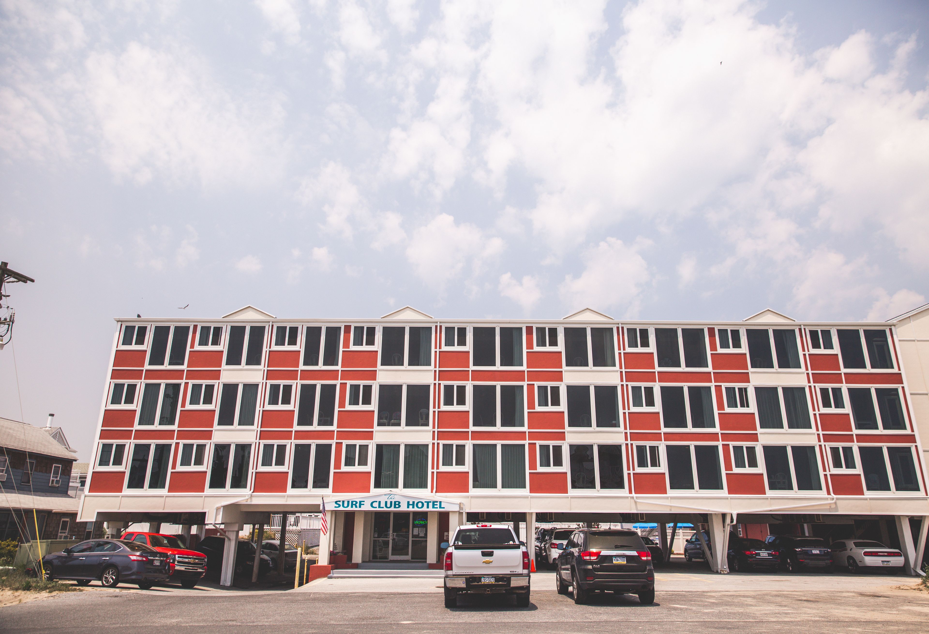 Un hôtel à plusieurs étages, avec une façade rouge et blanche, une enseigne indiquant « Surf Club Hotel » et plusieurs véhicules garés devant.