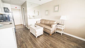 Una sala de estar moderna con un sofá color beige, una mesa de centro blanca y una mesita de noche con una lámpara. Se ve una zona de cocina al fondo.