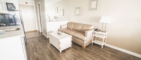 A modern living room with a beige sofa, a white coffee table, and a small side table with a lamp. A kitchen area is visible in the background.