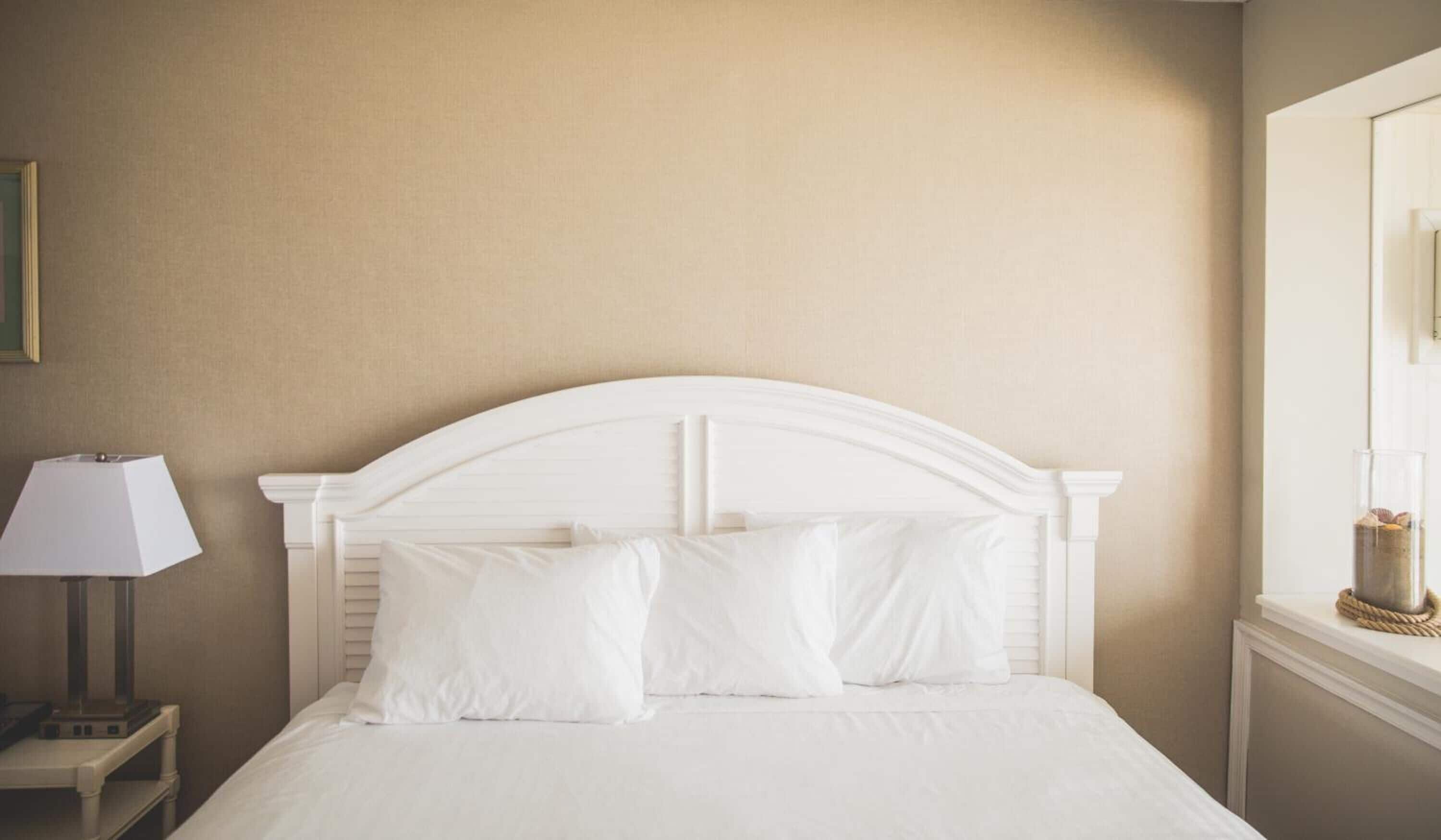 A neatly made bed with white bedding and pillows, a white nightstand with a lamp, and a window with a view of the outside.