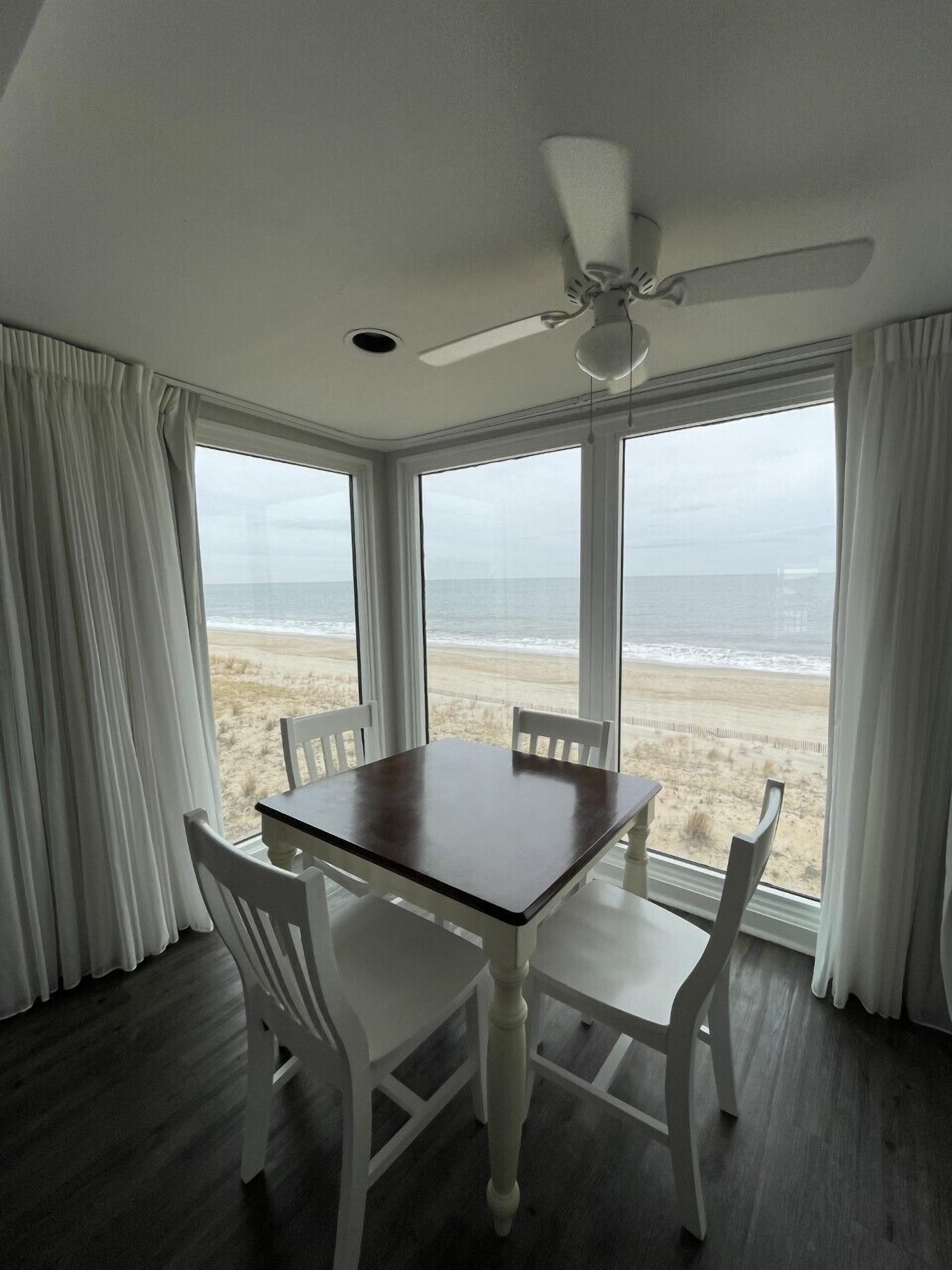 Una habitación con una mesa y cuatro sillas, ventanas amplias con vista a la playa y un ventilador de techo.