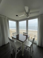 A room with a table and four chairs, large windows with a view of the beach, and a ceiling fan.