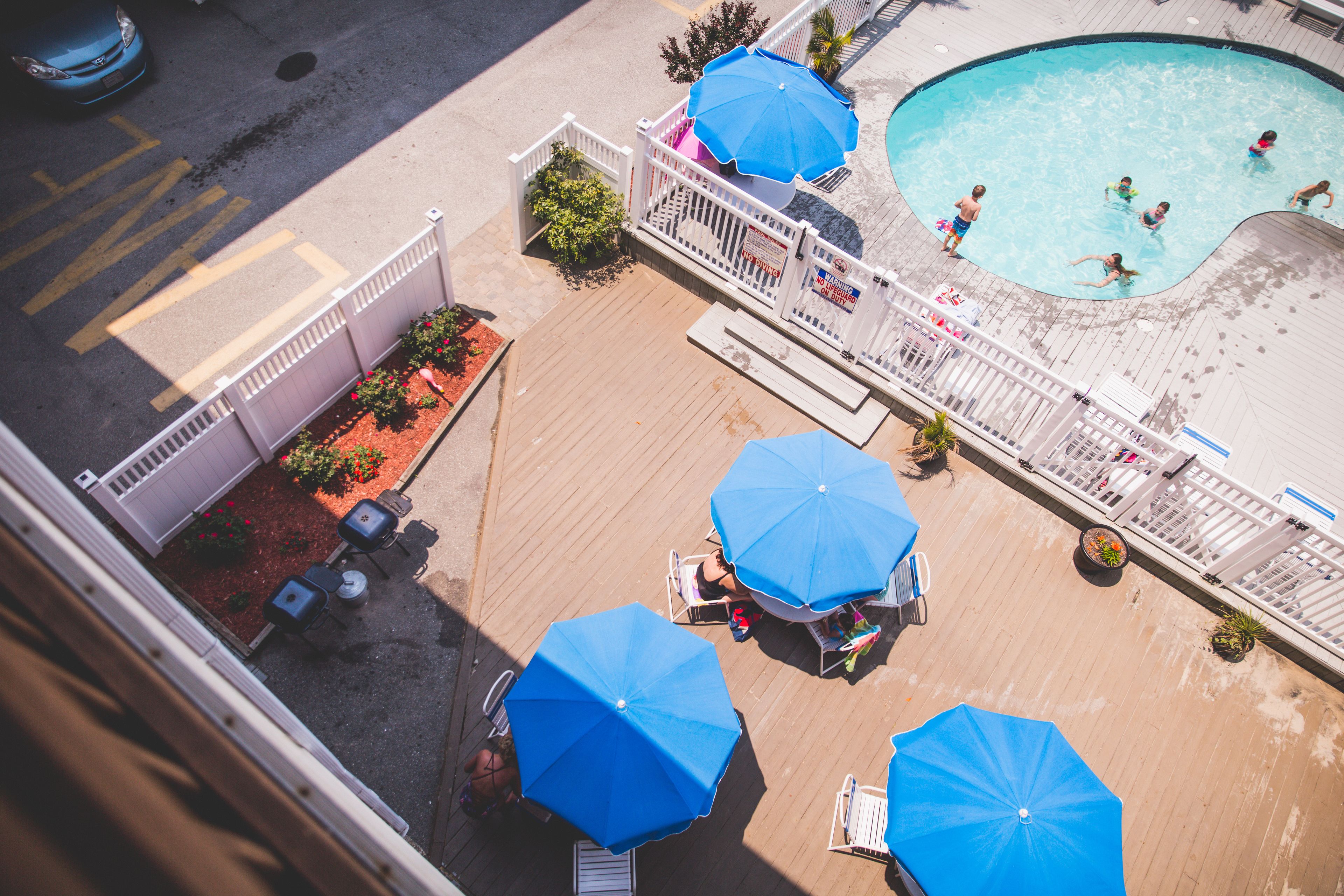 An aerial view of a swimming pool area with multiple blue umbrellas, a wooden deck, and a fenced poolside.