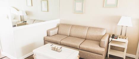 Un salon avec un canapé beige, un pouf blanc, une petite table d’appoint avec une lampe, et des tableaux encadrés accrochés au mur.