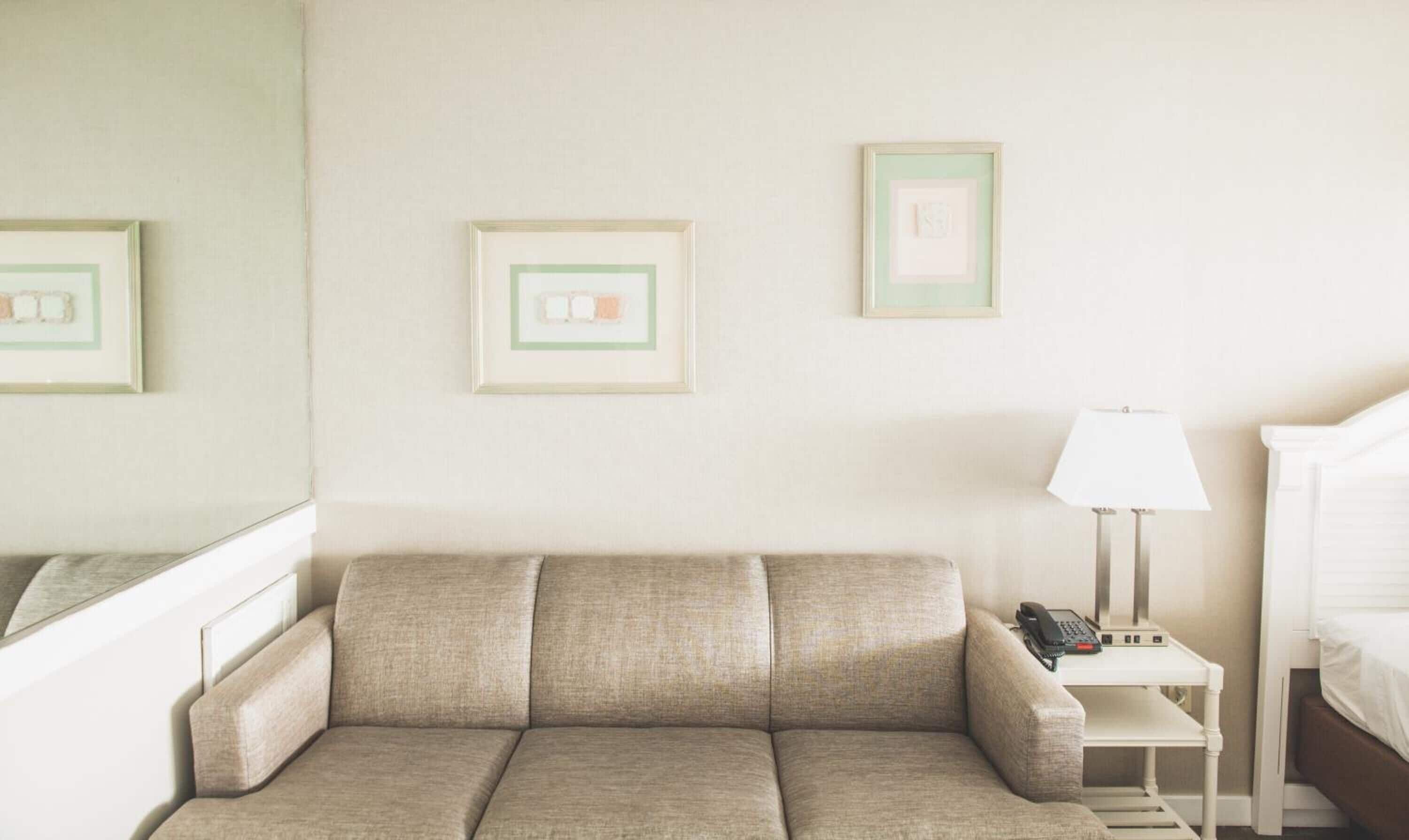Een hotelkamer met een beige bank, een klein nachtkastje met een lamp, een telefoon en een ingelijst foto op de muur.