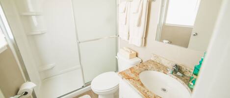 Baño con cabina de ducha de vidrio, inodoro, lavamanos con encimera de mármol y alfombra blanca.