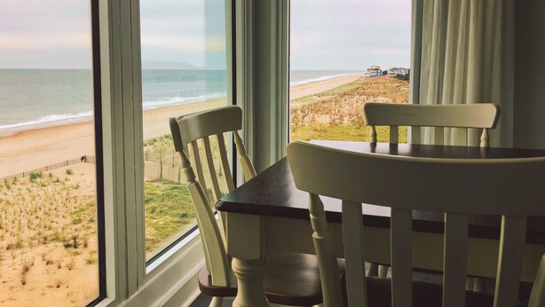 A room with a large window overlooking a beach, two wooden chairs, and a table. - Surf Club Oceanfront Hotel (Dewey Beach)