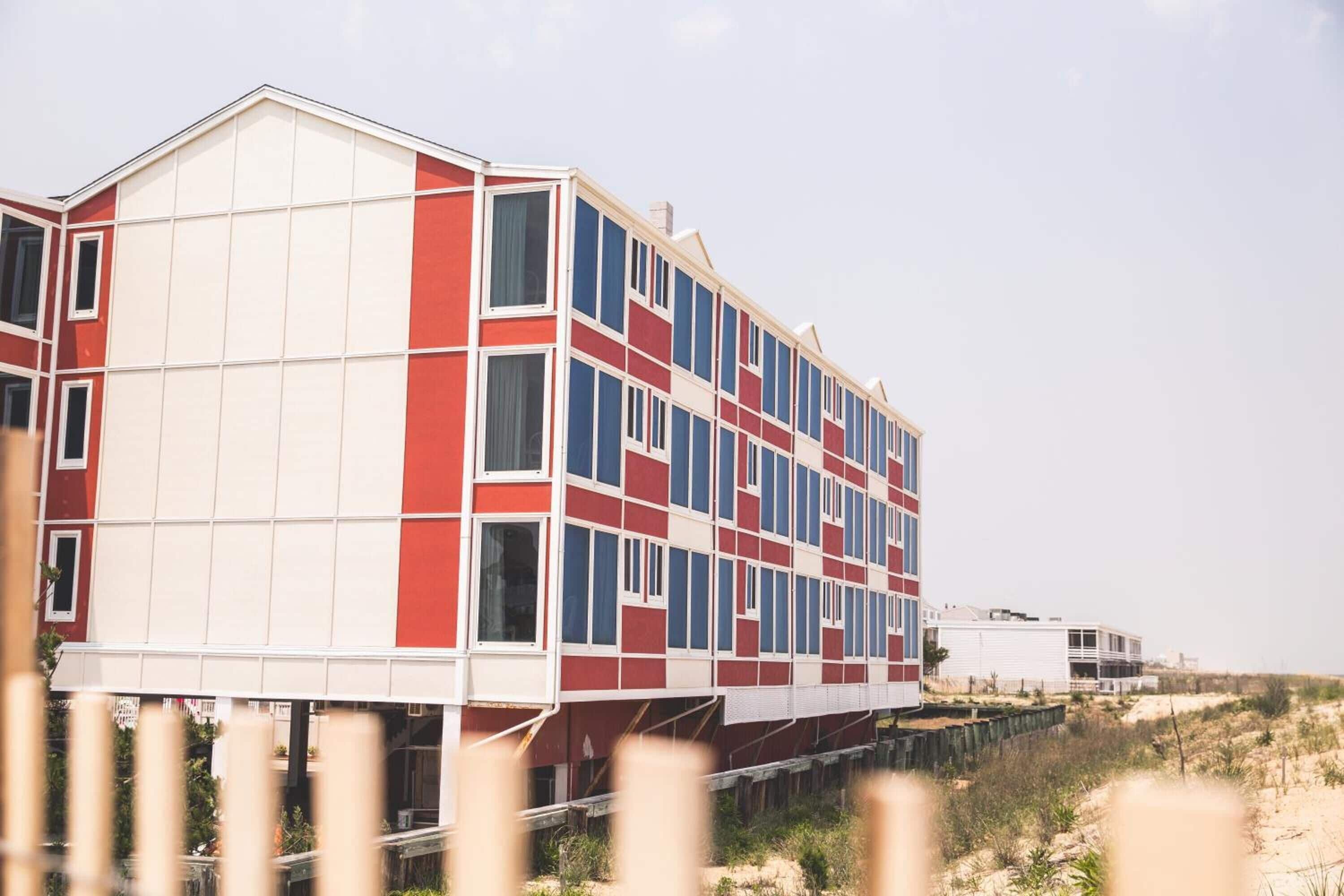 Un edificio de varios pisos con una fachada distintiva roja y blanca, situado cerca de una playa con dunas de arena.