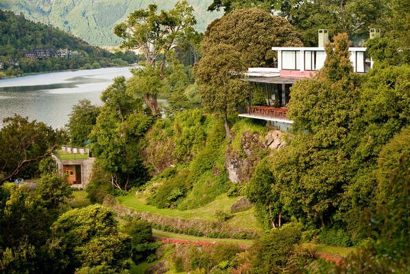 Une maison moderne à la façade en verre, donnant sur un lac, et entourée d’une végétation luxuriante et de collines.