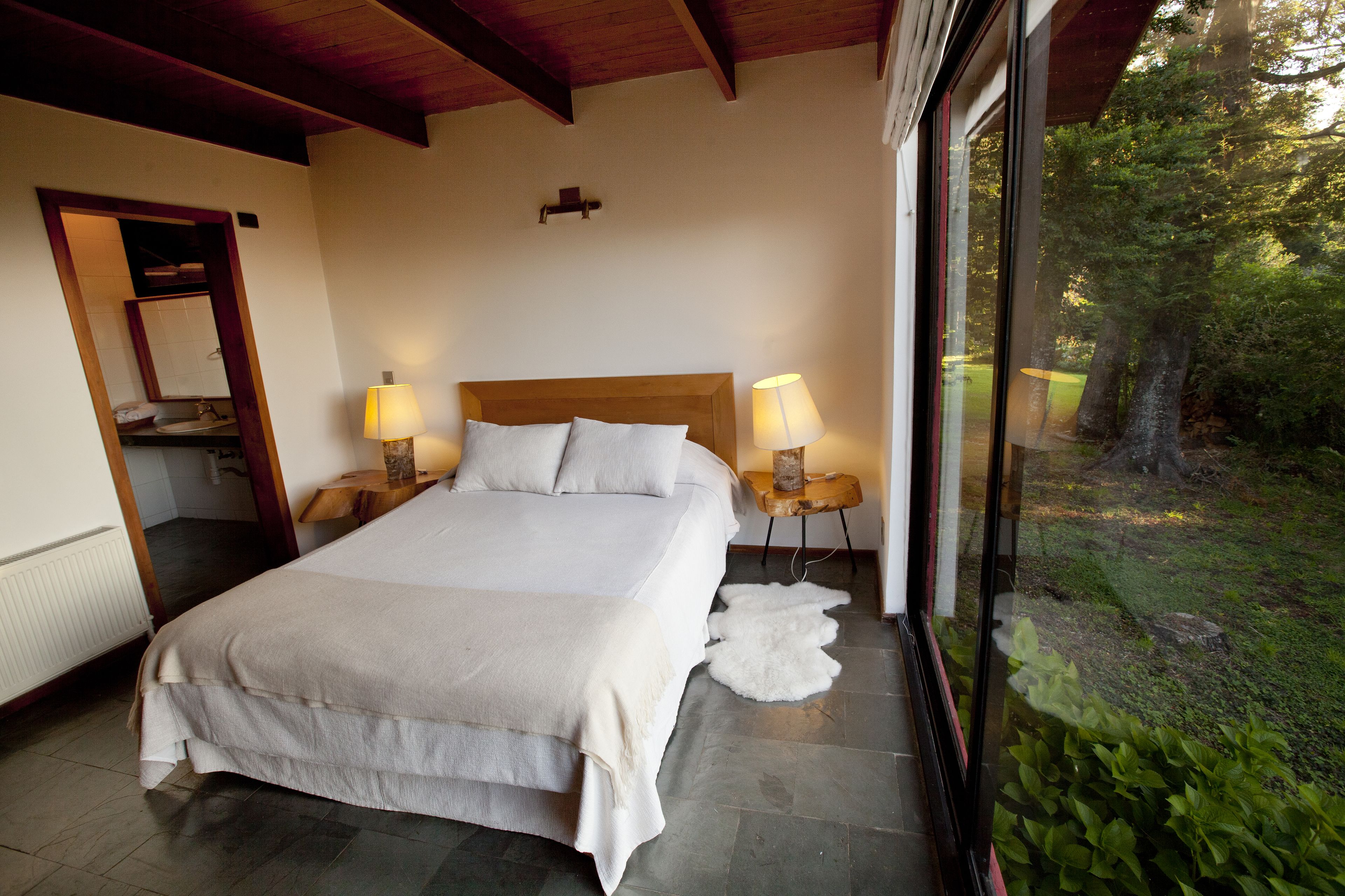 A bedroom with a large bed, two bedside lamps, a wooden headboard, and a view of the outdoors.