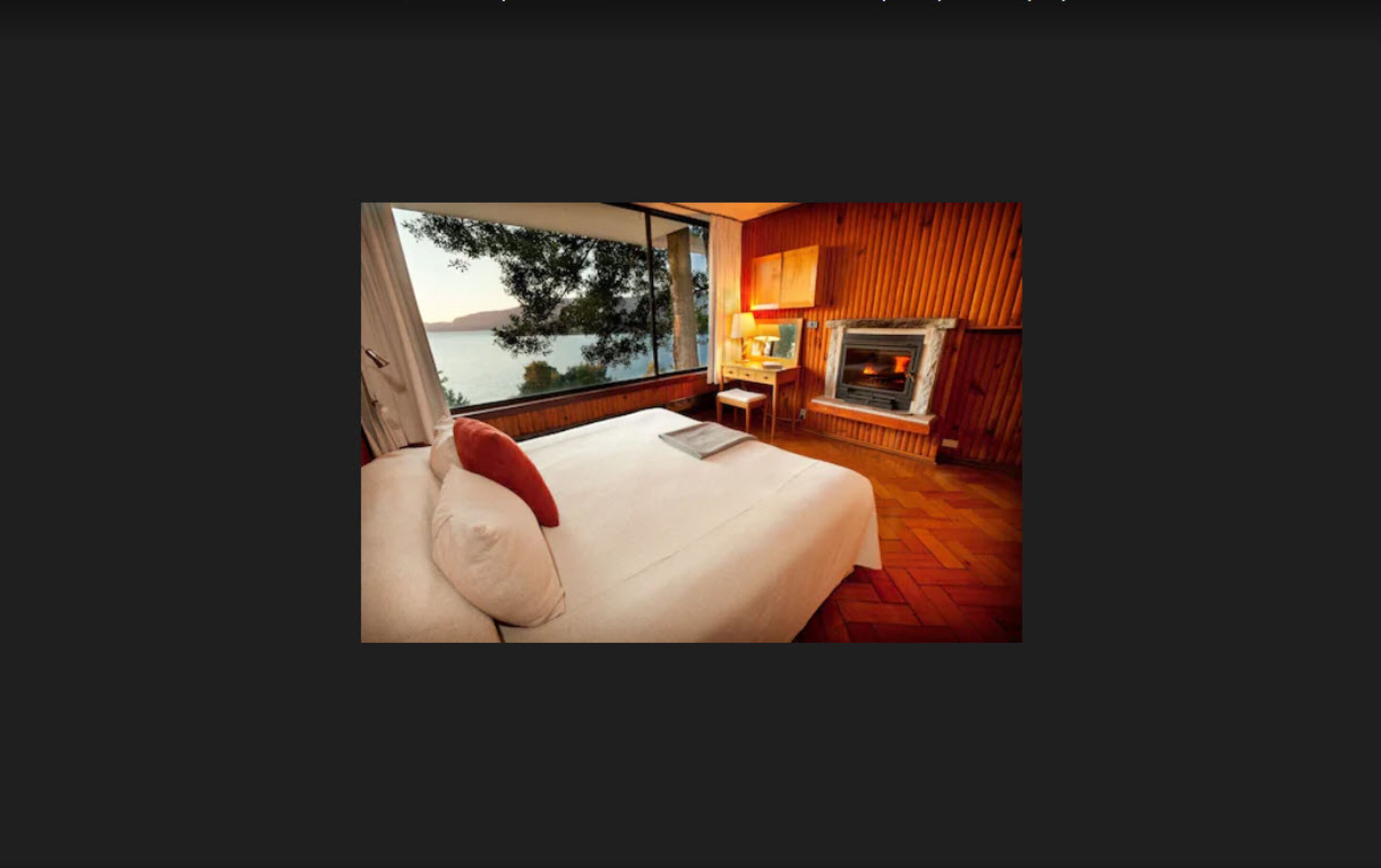 A bedroom with a large bed, a desk, a fireplace, and a view of a body of water.