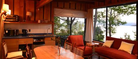 Interior acogedor de una cabaña con una mesa de madera, sillas, un sofá rojo y vistas a un lago a través de amplios ventanales.