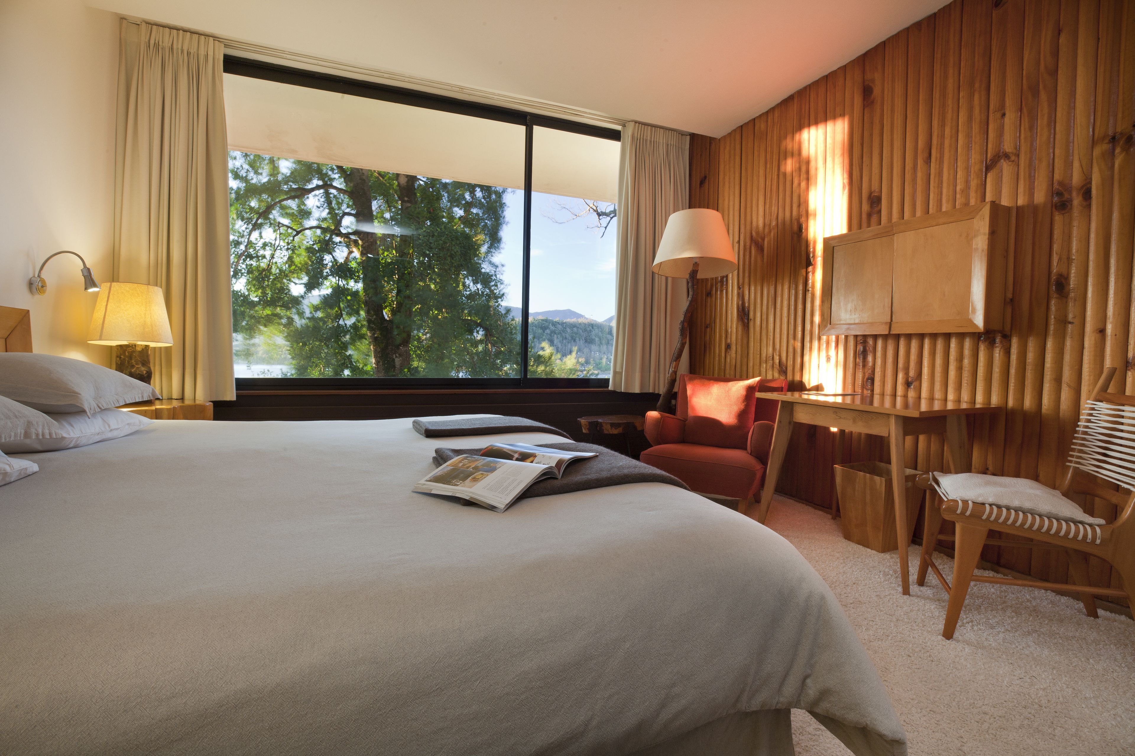 Quarto de hotel com uma cama grande, uma mesa de trabalho, uma cadeira e uma janela com vista para árvores e montanhas.
