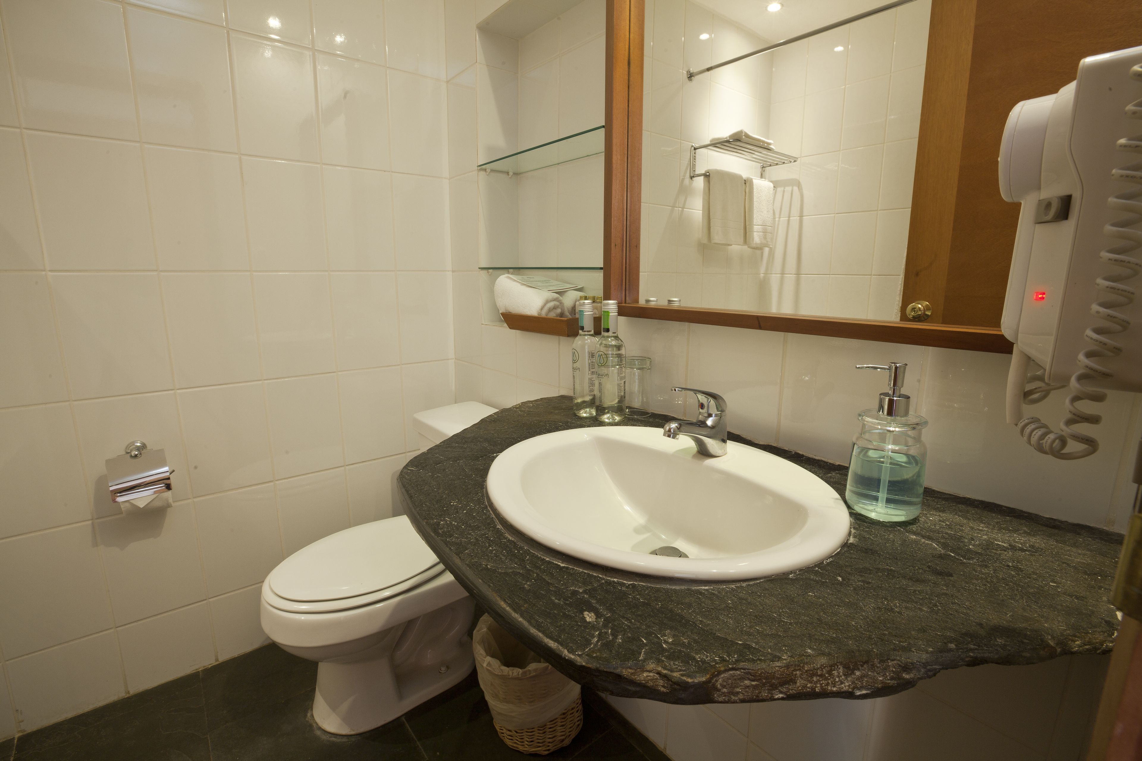 Une salle de bain avec des toilettes blanches, un lavabo doté d’un plan de travail en marbre, une étagère en verre au-dessus du lavabo et un miroir avec un porte-serviettes.
