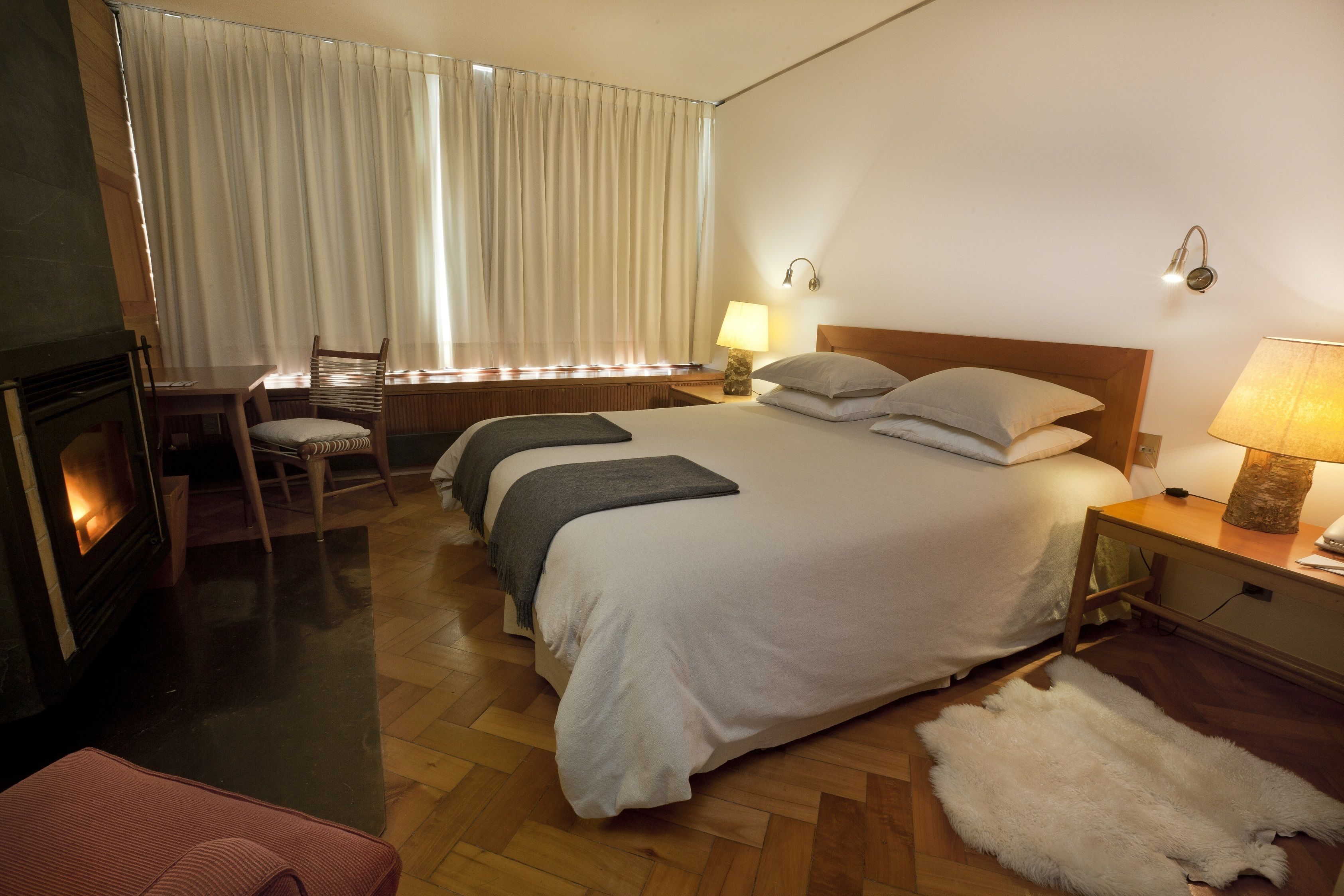 A hotel room with a large bed, a wooden headboard, a bedside table with a lamp, a chair, a fireplace, and a fur rug on the floor.