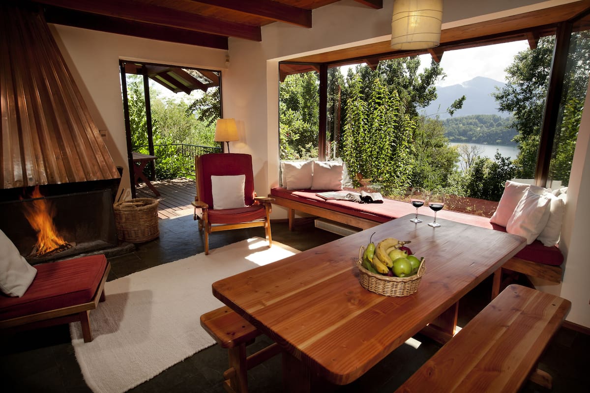Une pièce chaleureuse avec une cheminée, une table en bois sur laquelle est posé un panier de fruits, et une vue sur un lac et des montagnes.