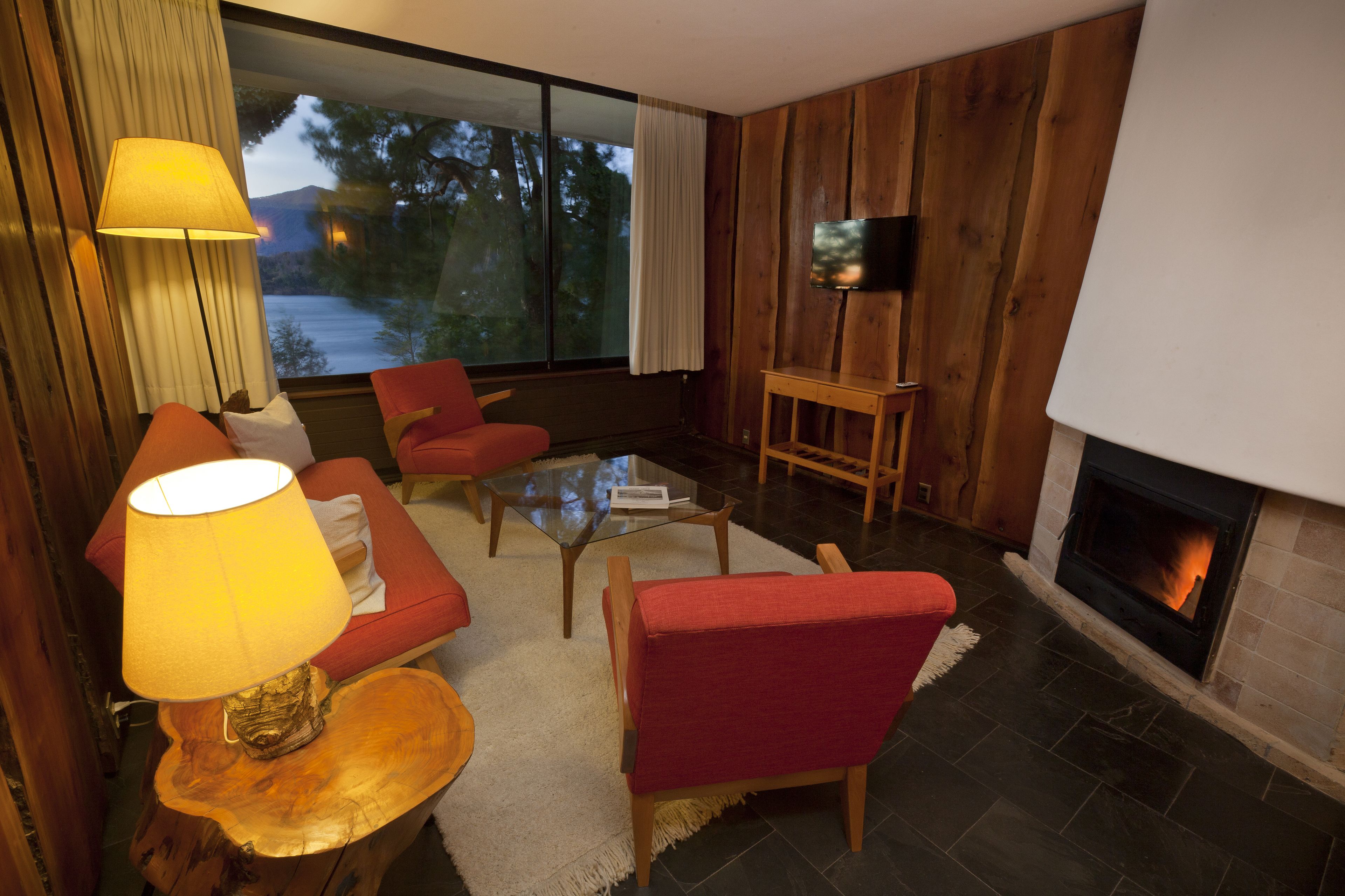 Een gezellige woonkamer met een open haard, een bank, een rode fauteuil, een glazen salontafel en een lamp.