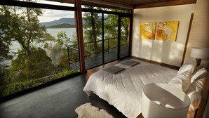 A bedroom with a large window overlooking a lake, a bed with white linens, and a view of trees and mountains. - Hotel Antumalal (Pucón)