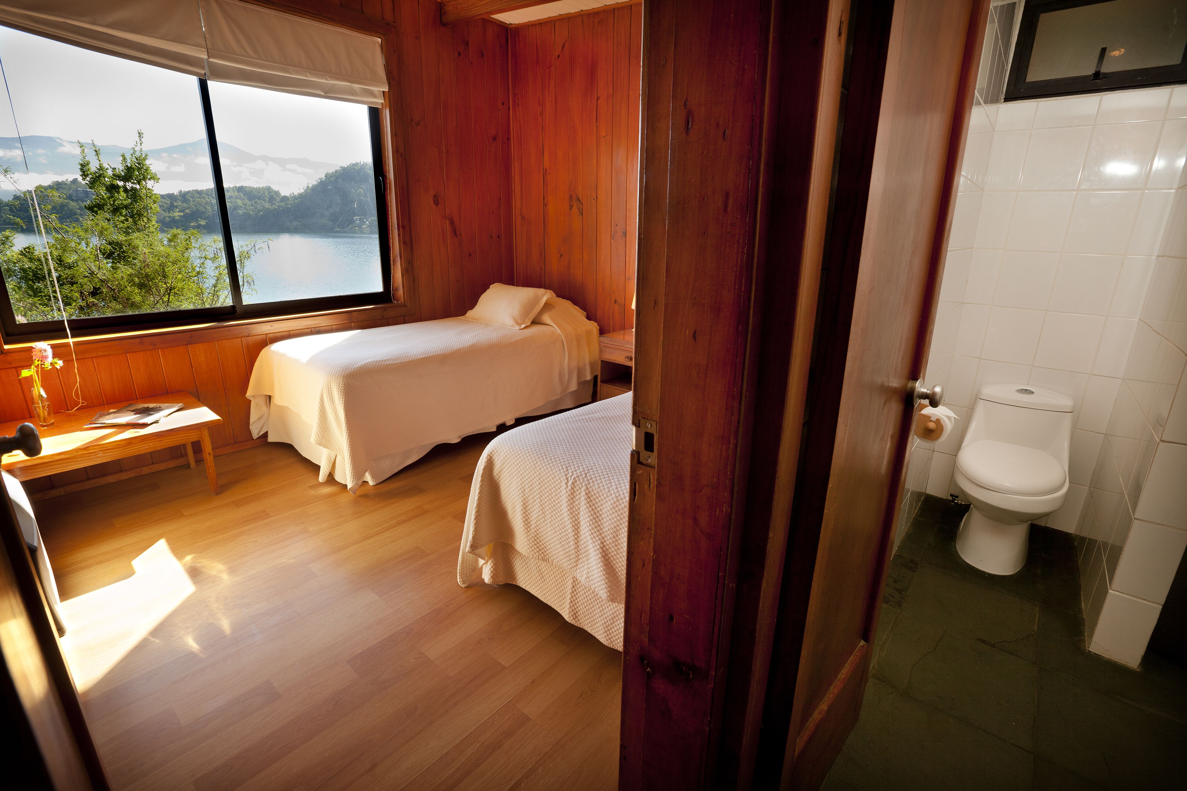Een kamer met twee bedden, een houten vloer, een raam met uitzicht op een meer en een badkamer met toilet.