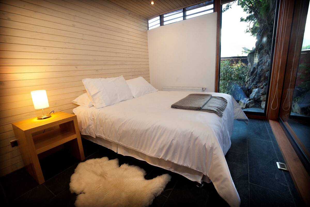 Une chambre avec un plafond en bois à lattes, un lit recouvert de draps blancs, une table de chevet avec une lampe, et une vue sur un jardin rocheux par une porte vitrée.