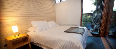 A bedroom with a wooden slatted ceiling, a bed with white linens, a bedside table with a lamp, and a view of a rocky garden through a glass door.