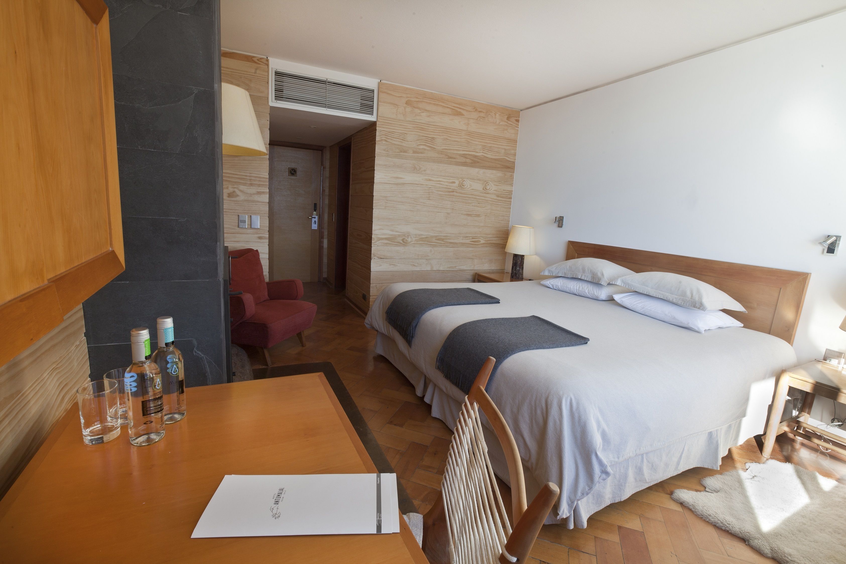 A hotel room with a bed, a wooden table with glasses and a menu, a chair, and a desk lamp.