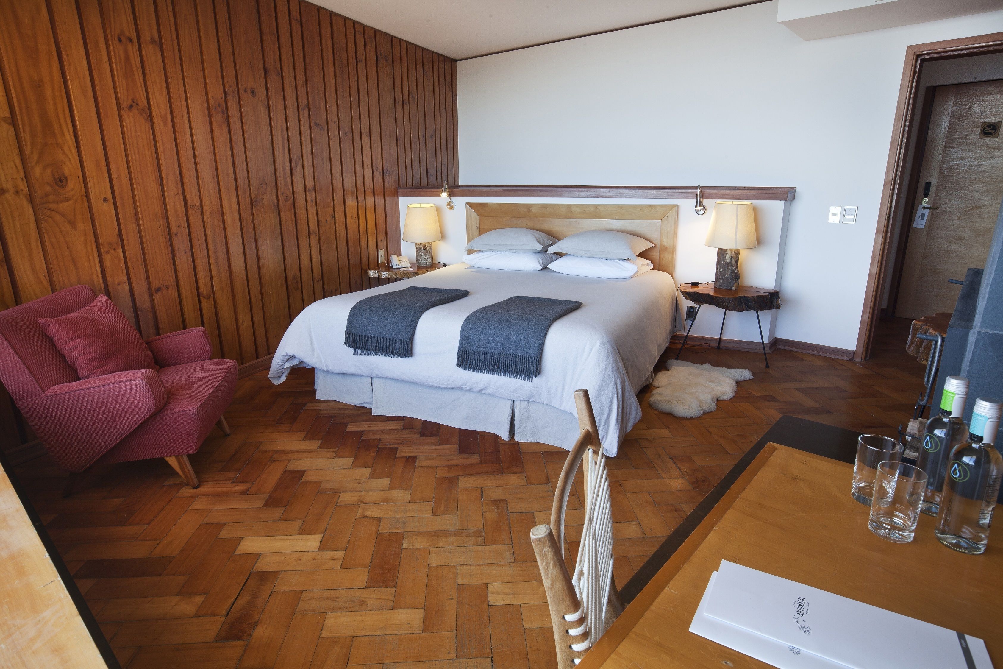 A hotel room with a wooden floor, a bed with white linens, a red chair, and a wooden headboard.